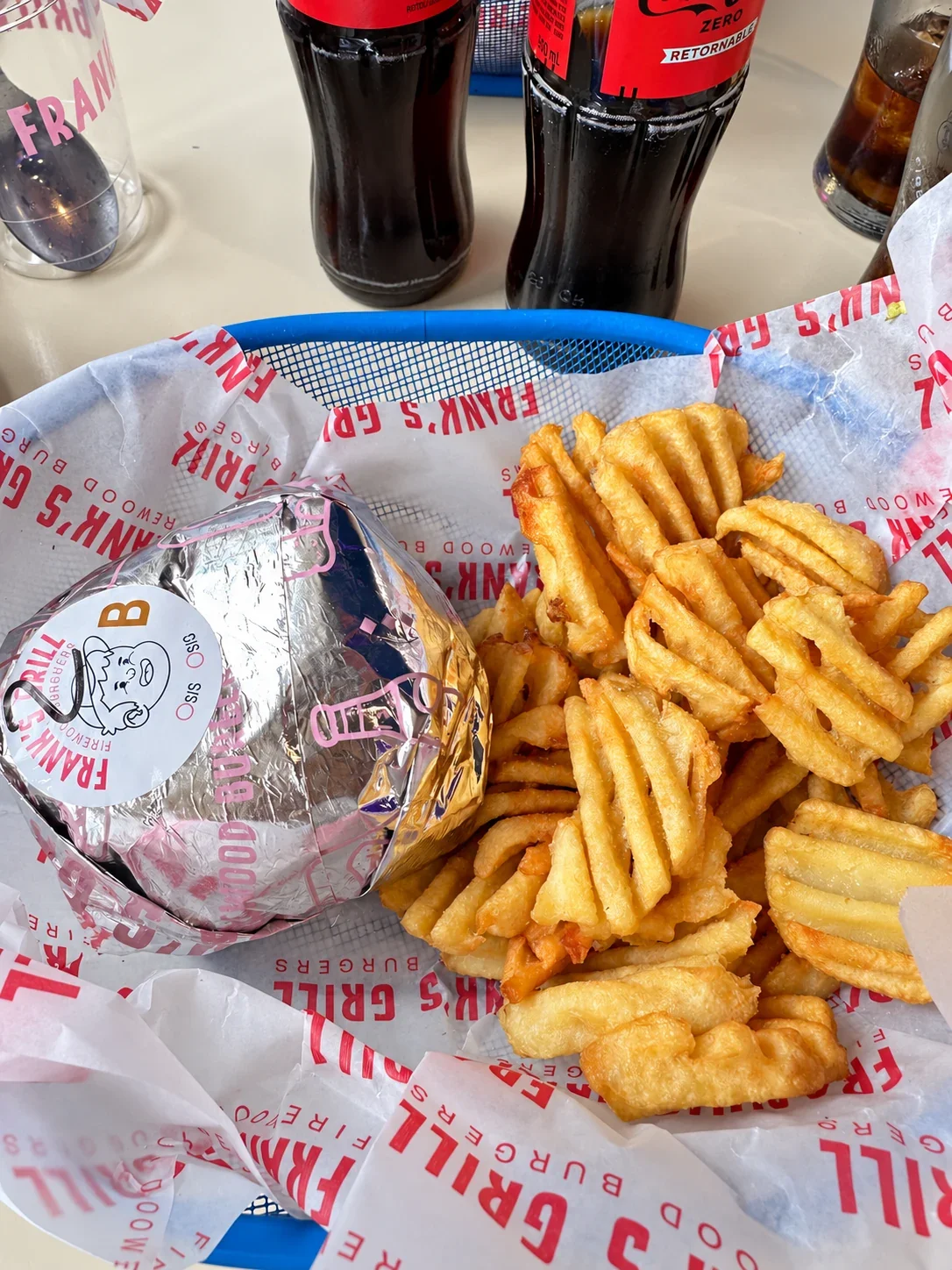 A Frank’s Grill burger wrapped in foil beside a serving of waffle fries in a blue basket