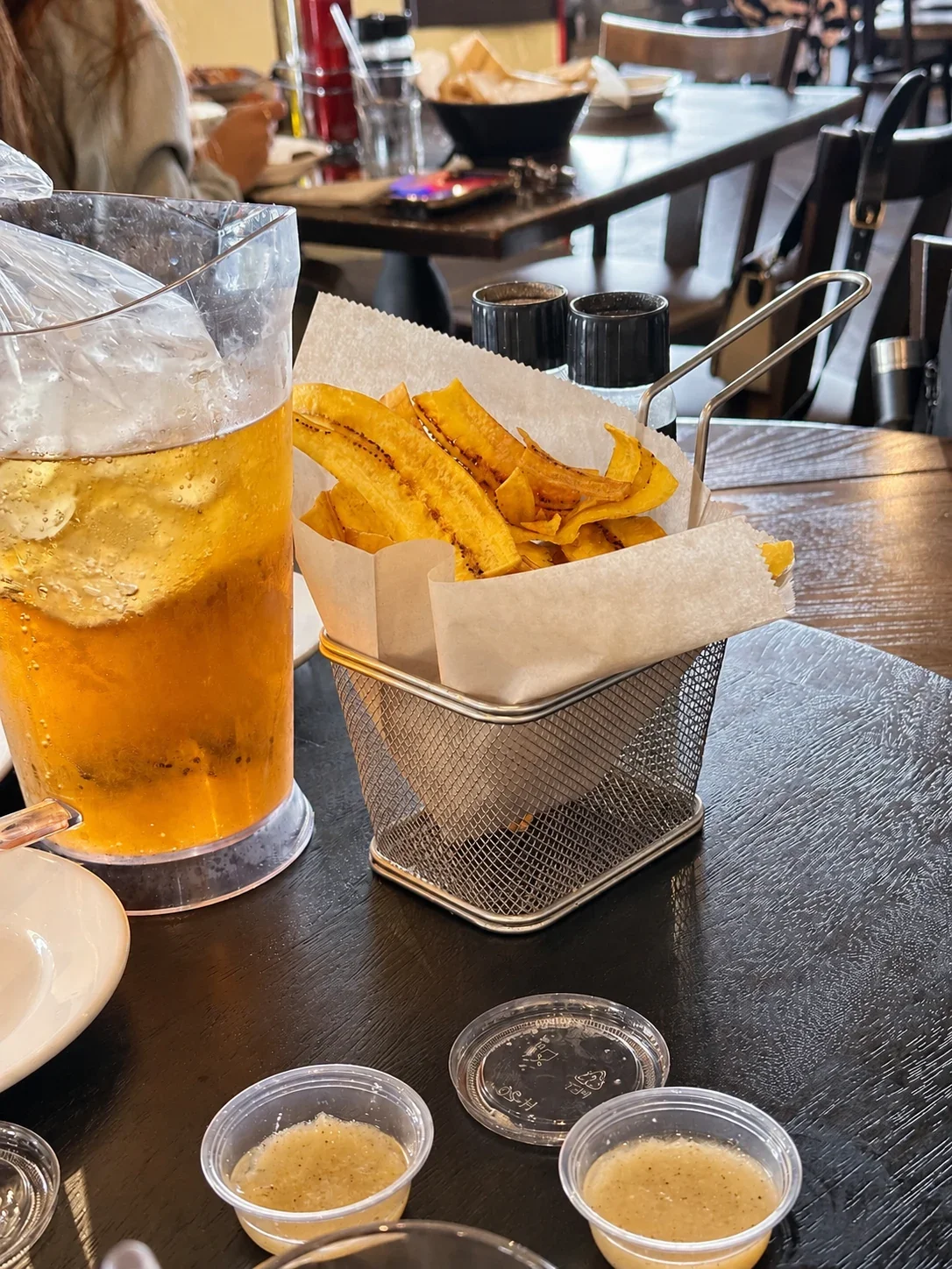 Thin fried plantain chips in a wire basket beside small cups of garlic mojo and a beer